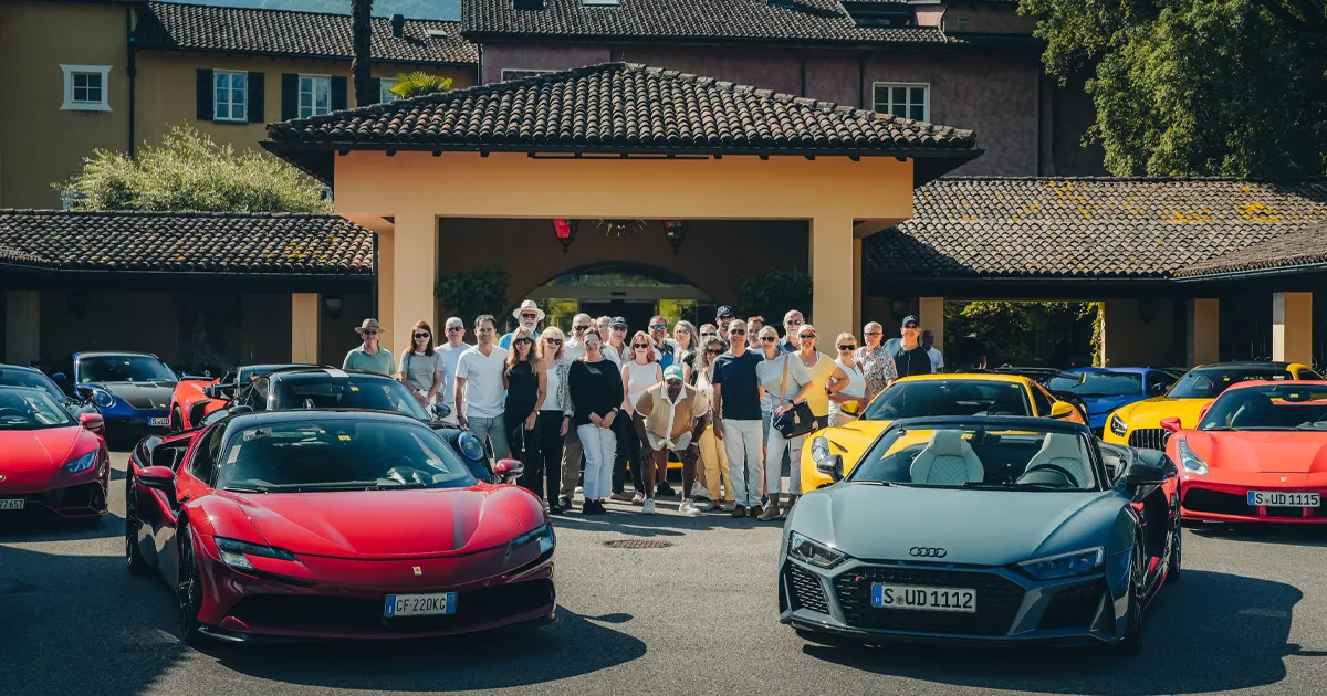 A group of people stand outside a luxury villa flanked by Ultimate Driving Tours supercars