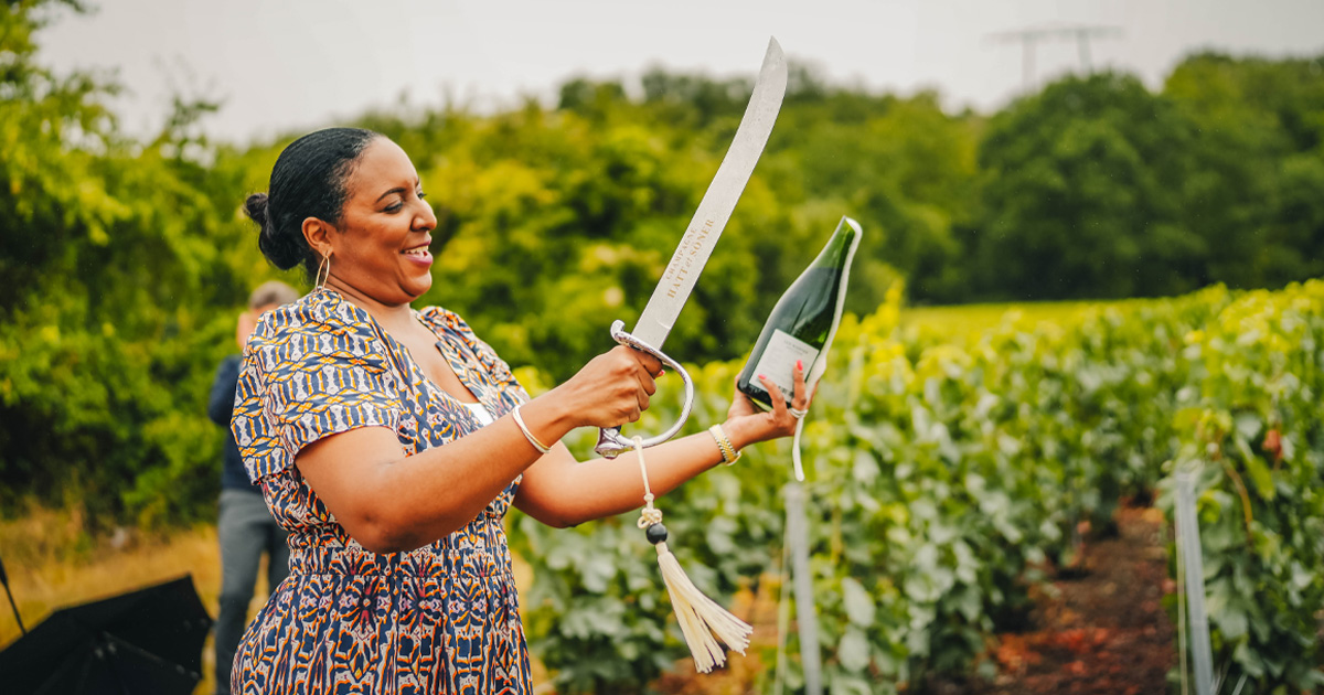 A lady smiles as she removes the cork from a champagne bottle using a traditional sabre