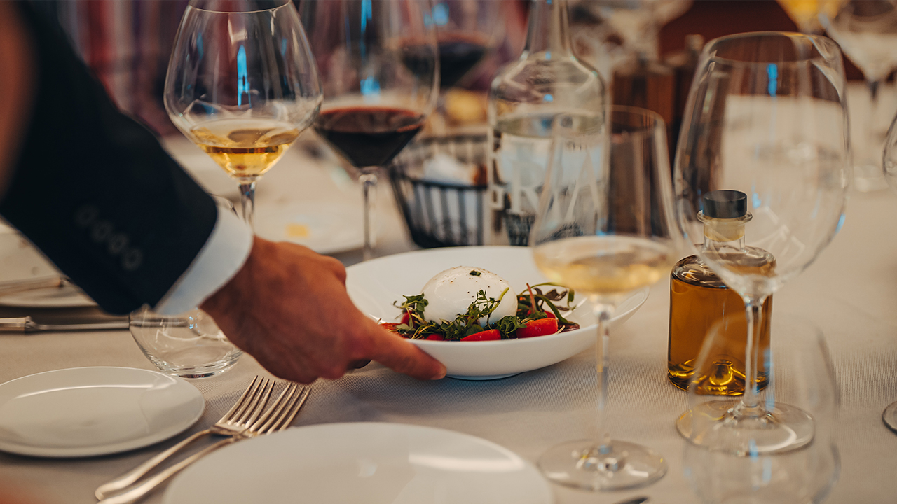A close up of a waiter presenting an elegant salad to a table adorned with white tablecloth and glasses filled with white wine