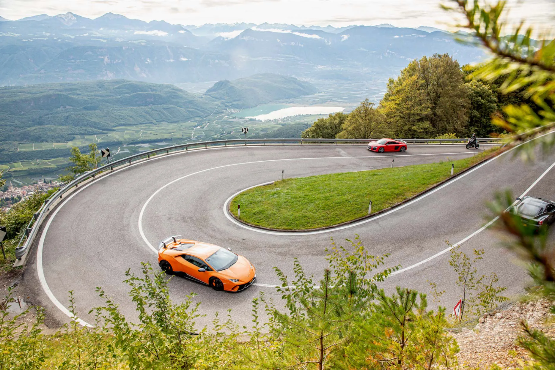 three supercars drive up a steep road with a valley and a mountain range in the background