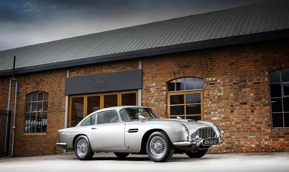 An immaculate example of the Aston Martin DB5 sits outside a brick building bearing the Aston Martin insignia and the word ‘heritage’