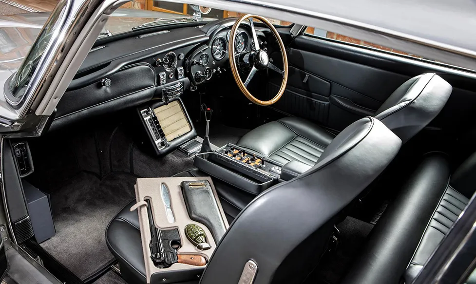 The interior of a James Bond Aston Martin DB5 shows special modifications to the black leather trim, including a box containing a pistol and a hand grenade