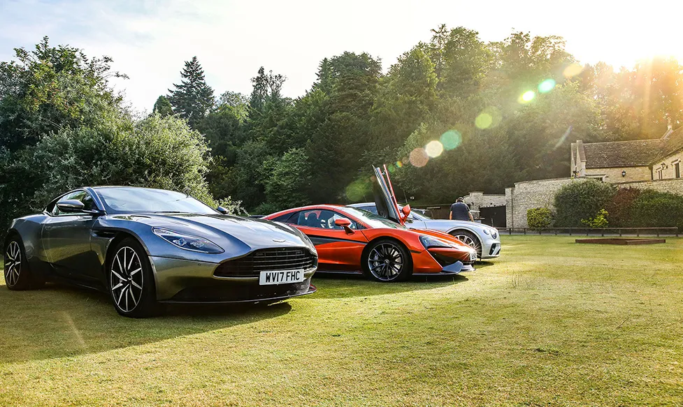 A brand new Aston Martin DBS belonging to the Ultimate Driving Tours fleet sits next to a red McLaren on a grassy lawn in the afternoon sun