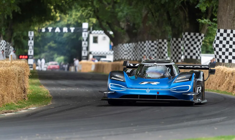 A blue Volskwagen racing car prototype navigates a sweeping corner at Goodwood Festival of Speed with the start line visible in the rear distance