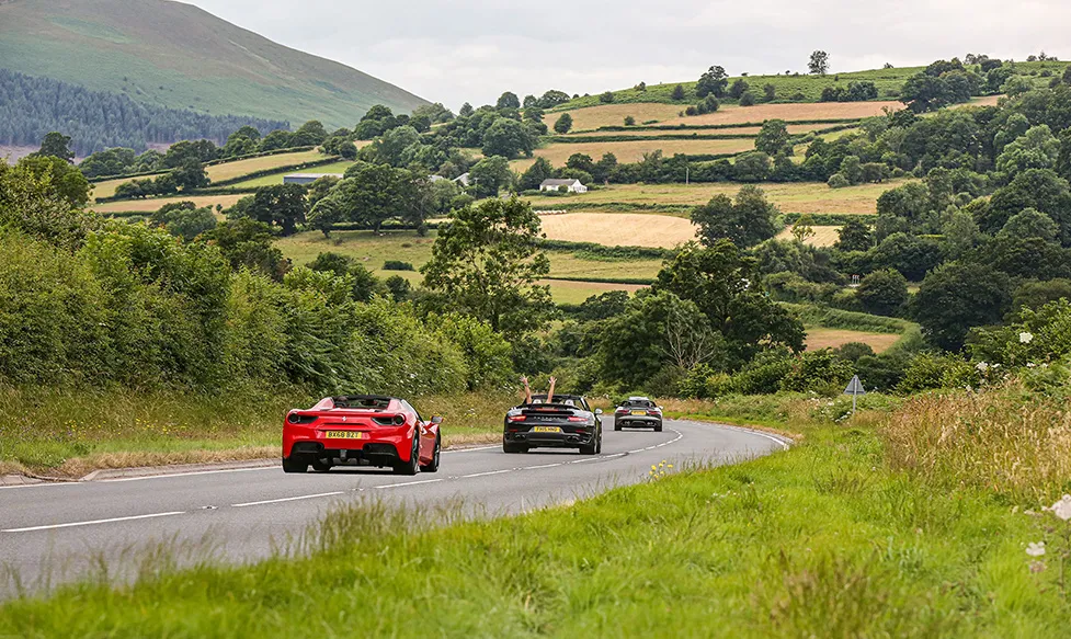 Three supercars approach a corner in the English countryside on a fine day. The passenger of the middle car has their hands raised joyfully in the air
