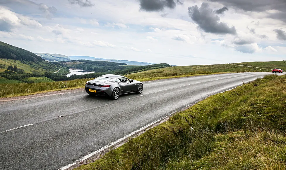 A gunmetal grey, late-model Aston Martin DBS crests a hill in the English countryside on an Ultimate Driving Tours supercar holiday