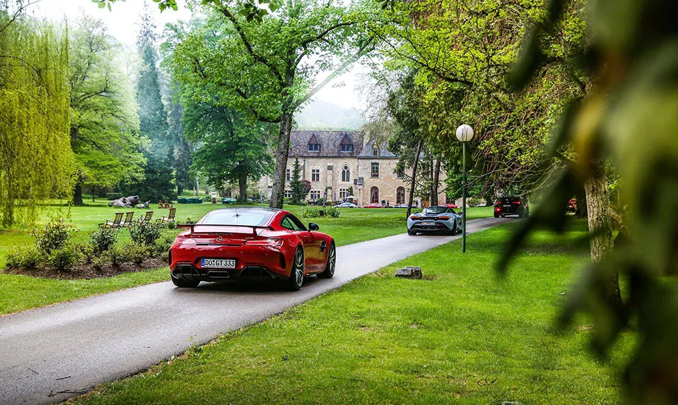 A red Mercedes GTR follows a McLaren up a driveway towards a regal country manor