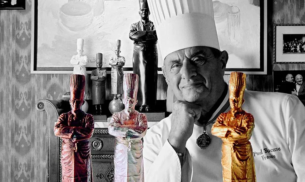 A black and white shot of legendary French chef, Paul Bocuse, standing behind some of his culinary awards