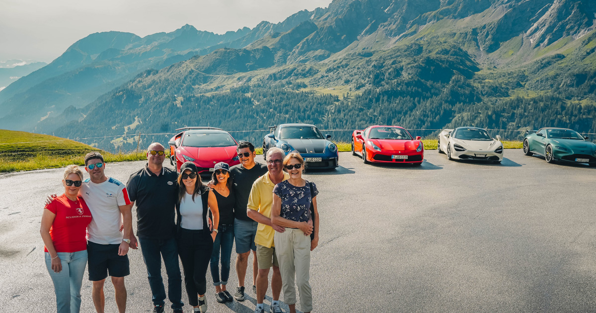 A small group of supercar tour guests pose in front of a neatly arranged fleet of supercars