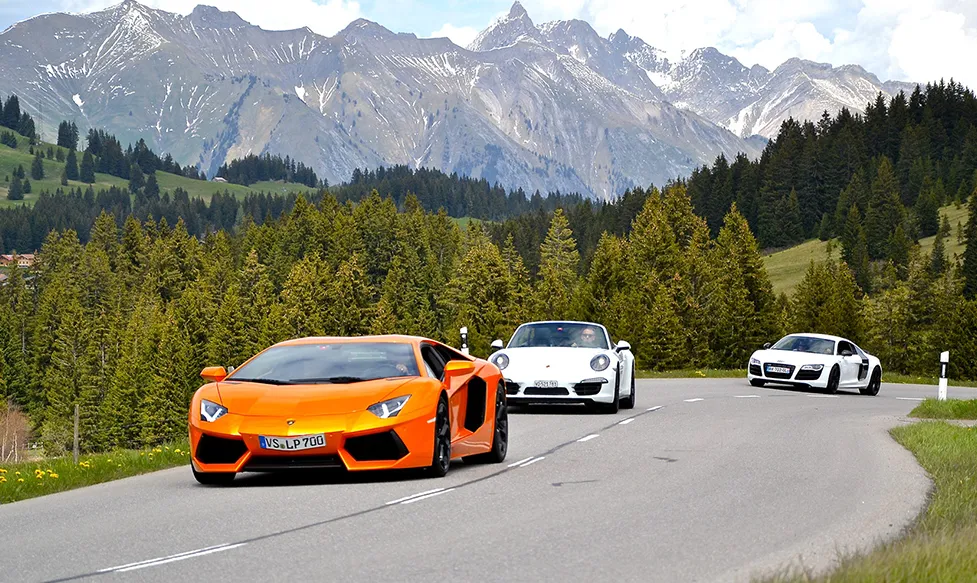 An orange Lamborghini Aventador drives a road ahead of a white Porsche 911 and a white Audi R8 against a dramatic mountain backdrop