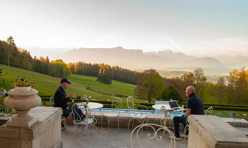 Two men sit on a large stone balcony conversing over their laptops as they take in a view across sweeping green valleys in the evening sun.