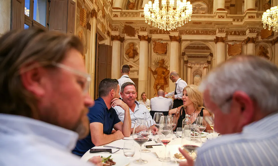 A group of people are talking around a large dinner table enjoying a meal in an ornate European dining room during their luxury tour