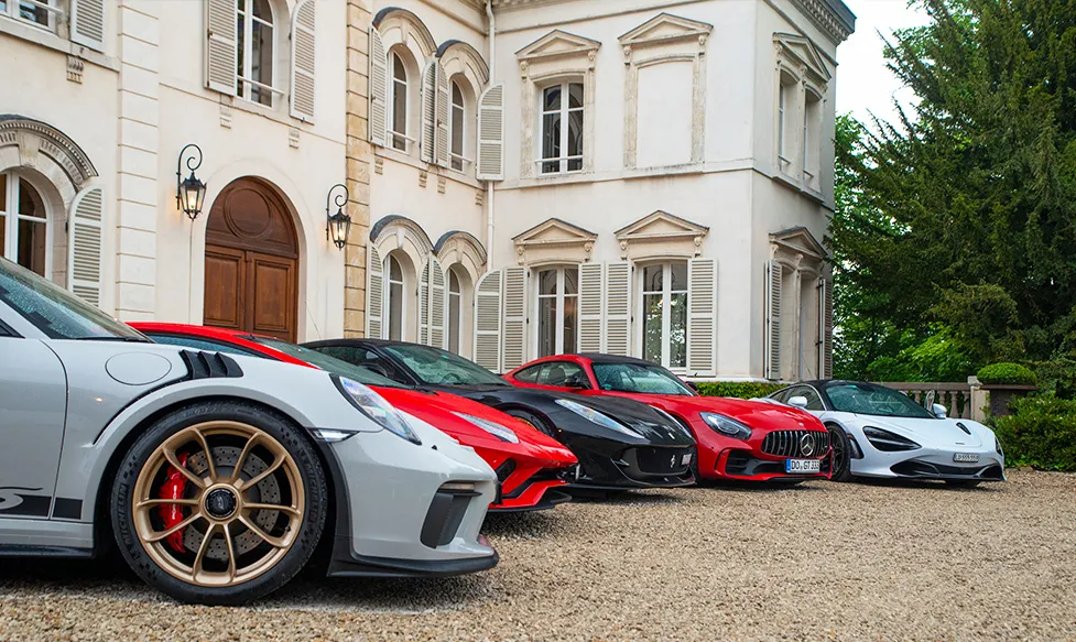 A selection of supercars sit on the gravel outside an elegant white stone mansion in France during an Ultimate Driving Tours luxury holiday.