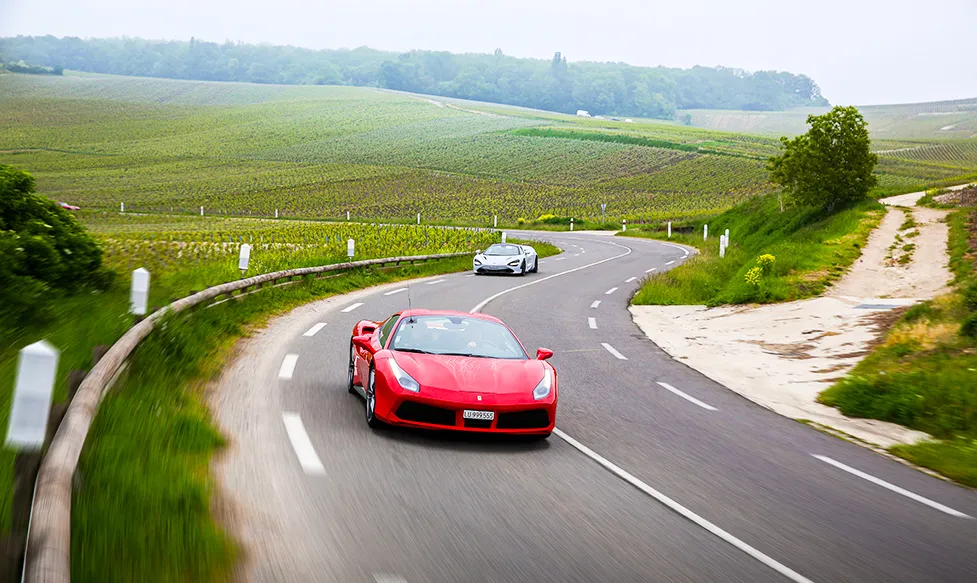 A red Ferrari leads a grey McLaren through a fast-paced section of road in Champagne, France