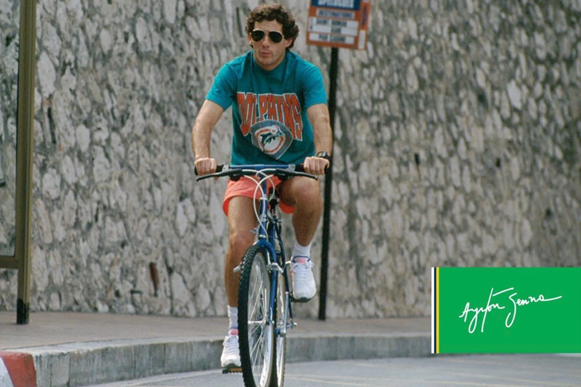 Ayrton Senna rides a bicycle wearing a Miami Dolphins t-shirt.