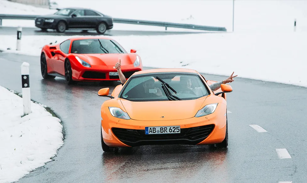 An orange McLaren is driven on a snowy road. The driver and passenger gesture with thumbs up and a peace sign